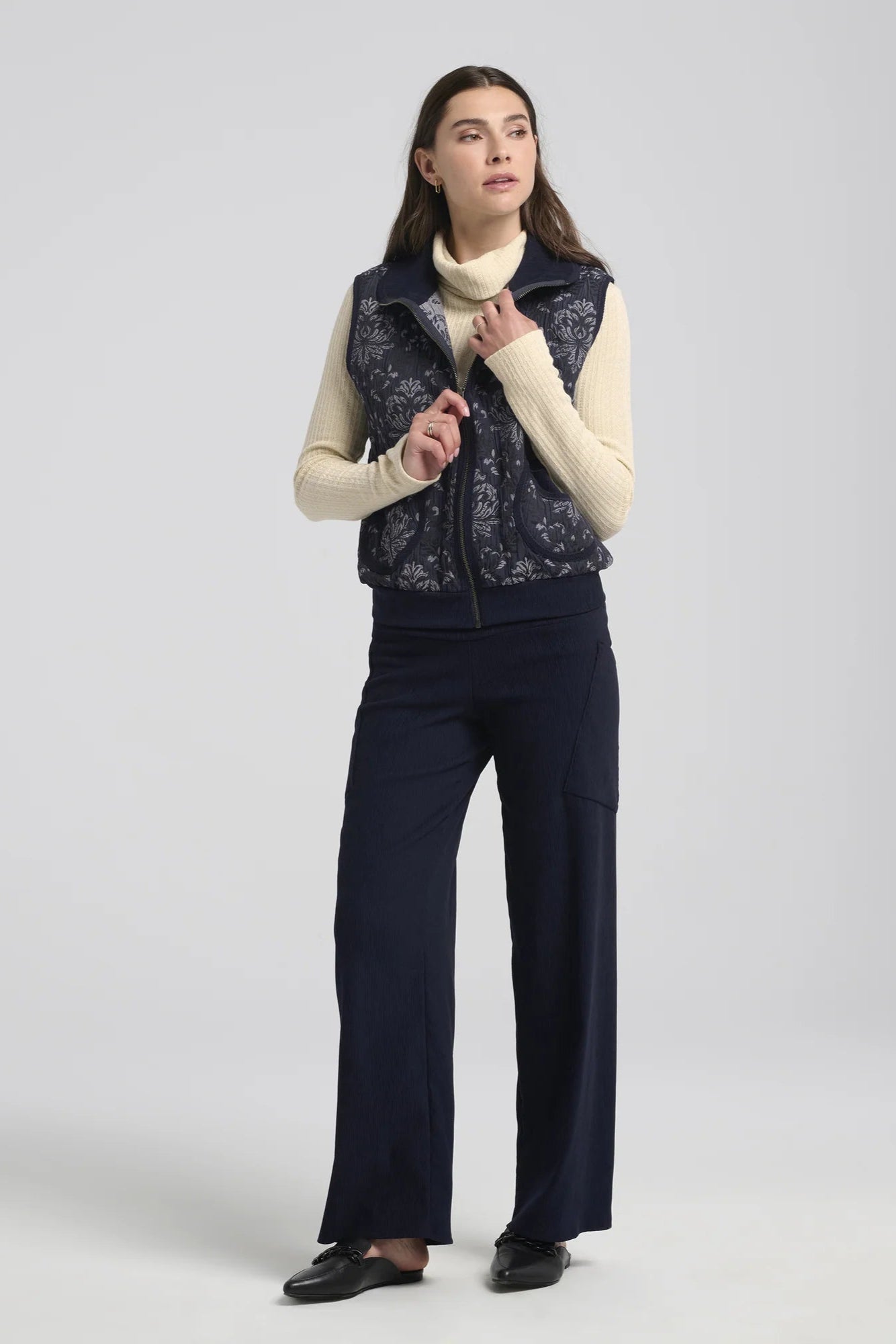 A woman wearing the Hermione Pants by Kollontai in Navy, stretchy corduroy pants with a wide pull-one waistband, wide legs, and angled patch pockets. She is wearing them with a cream sweater and a vest and standing in front of a grey background. 