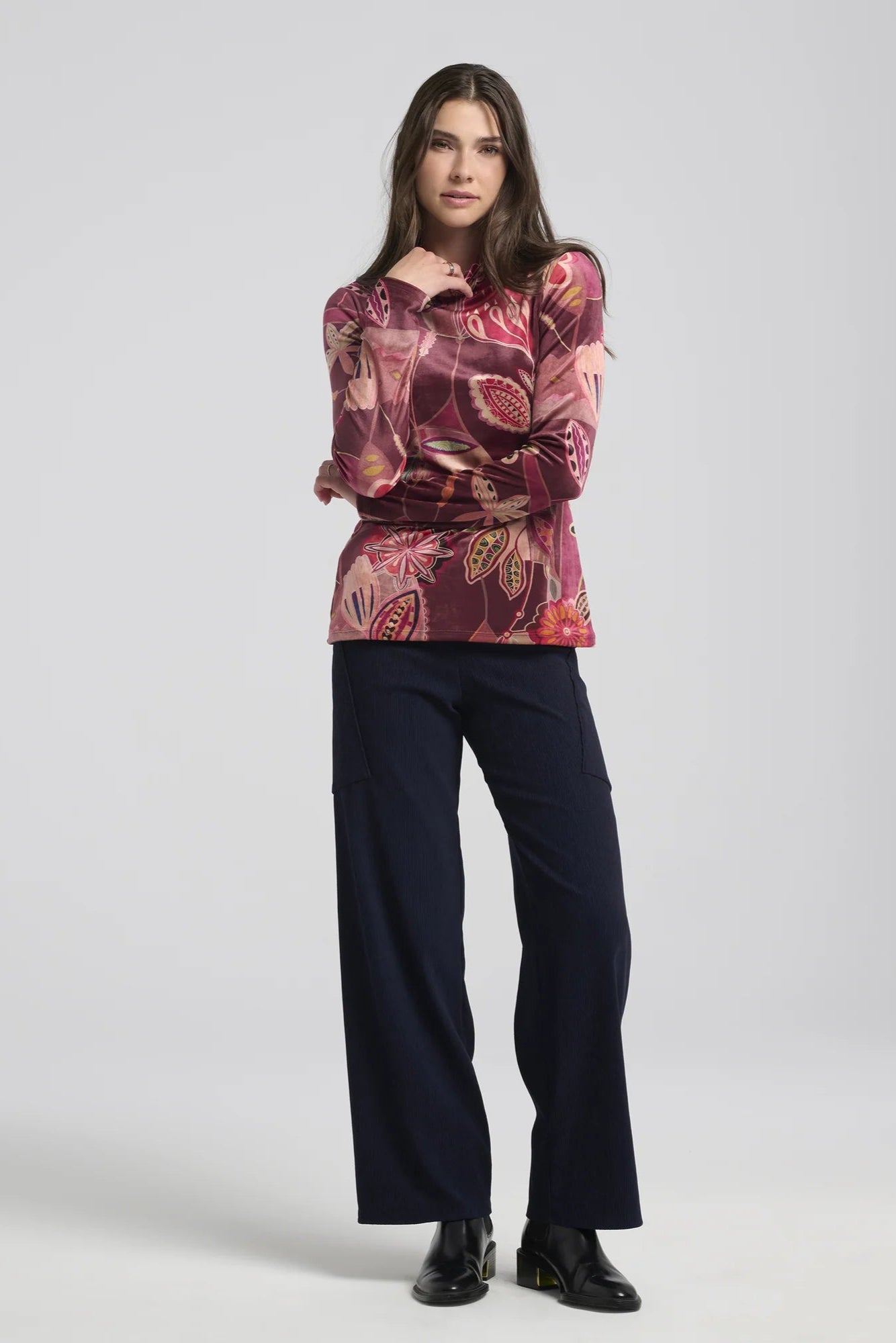 A woman wearing the Hermione Pants by Kollontai in Navy, stretchy corduroy pants with a wide pull-one waistband, wide legs, and angled patch pockets. She is wearing them with patterned burgundy top and standing in front of a grey background. 