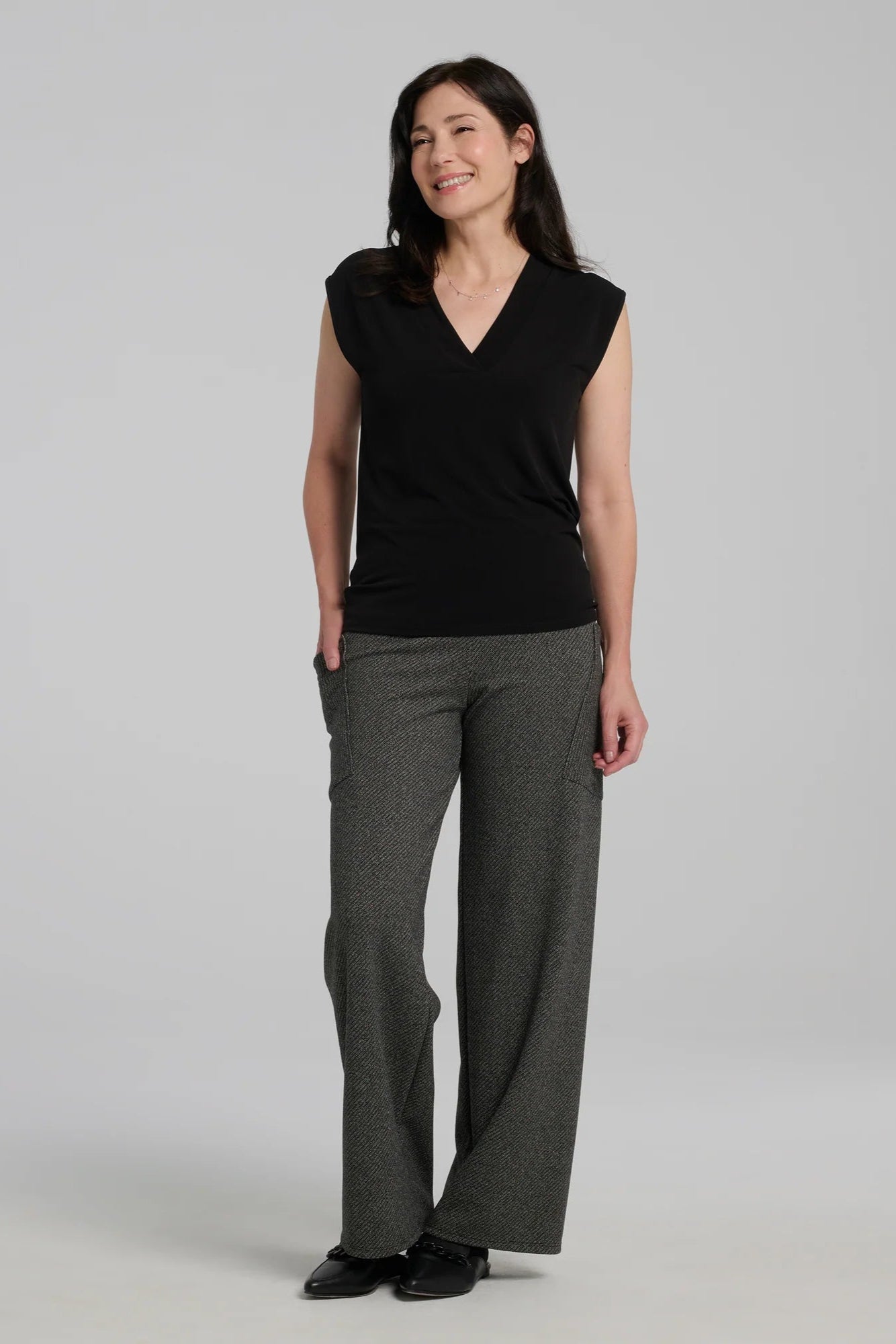A woman wearing the Susan Pants by Kollontai in Charcoal, wide pants with a wide pull on waistband and angled patch pockets. She is wearing them with a black top and standing in front of a grey background. 