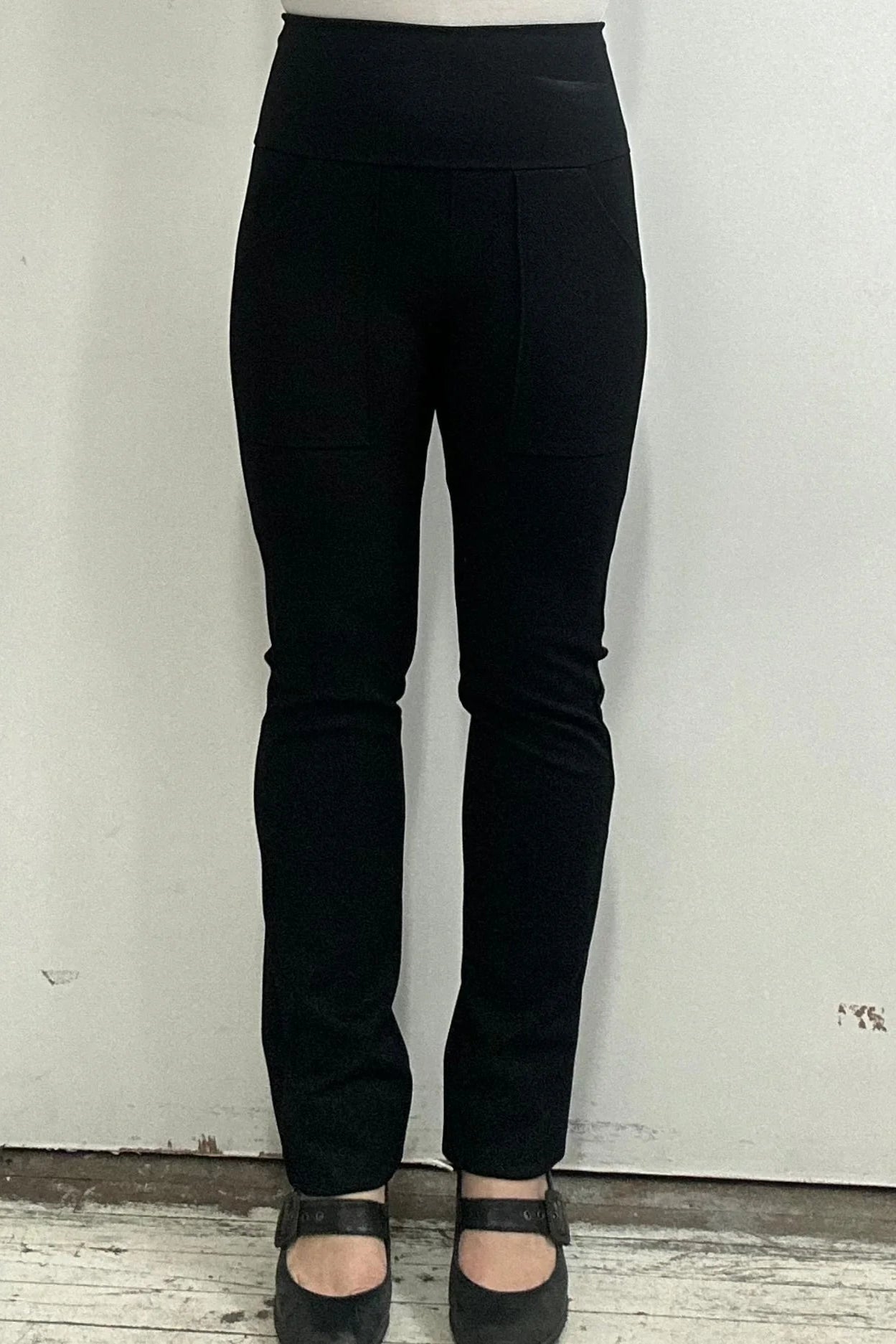 Waist down view of a woman wearing the Bari Pants by Kollontai, black pants with a slim fit, wide pull-on waistband, and front patch pockets, in front of a white wall. 