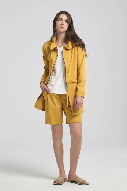 A woman wearing the Bronwen Jacket by Kollontai in Mustard, a mid-thigh length jacket with drawstrings at the waist and hood and large patch  pockets. She is wearing it with matching shorts and standing in front of a white background. 