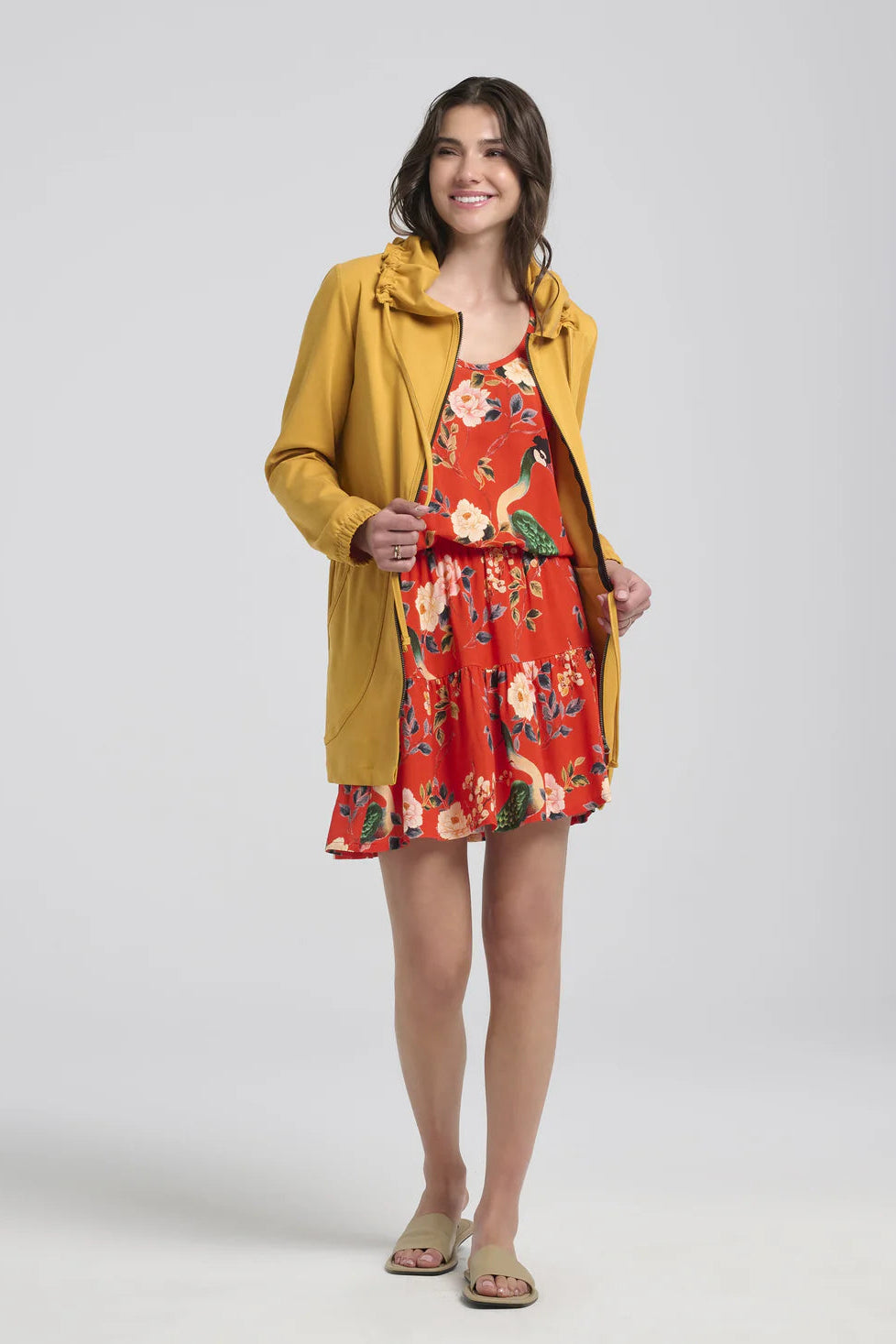 A woman wearing the Bronwen Jacket by Kollontai in Mustard, a mid-thigh length jacket with drawstrings at the waist and hood and large patch  pockets. She is wearing it a floral romper and standing in front of a white background. 