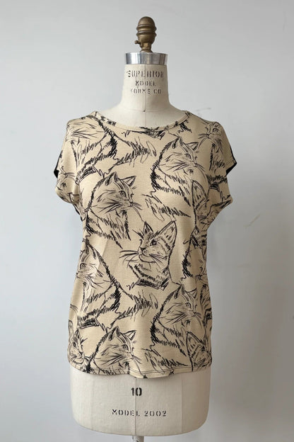 The Arty T-Shirt by Kollontai in Cats/Beige print, with a round neck, short extended sleeves, and a slight hi-low hemline, and a solid black back, is shown on a mannequin against a grey background. 