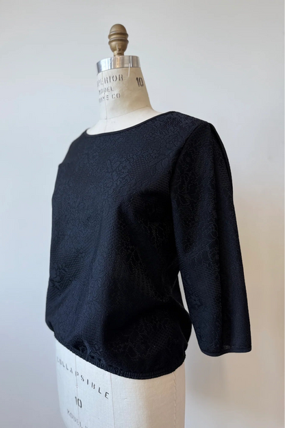 The Celestine Blouse By Kollontai in Black, featuring black satin overlaid with black lace, is shown on a mannequin in front of a white background. It has a cropped silhouette with an elastic waist, and 3/4 sleeves. 