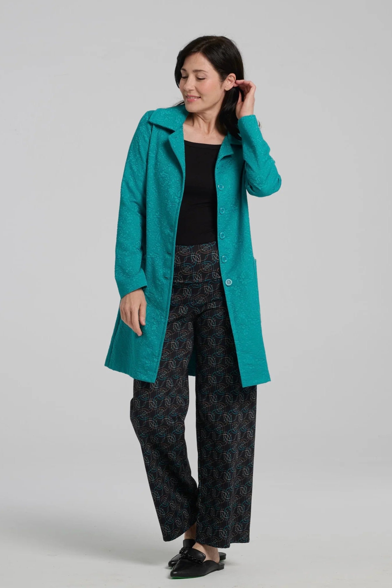 A woman wearing the Emmanuelle Coat by Kollontai in Turquoise jacquard with the Elma Pants. She is standing in front of a white background. 