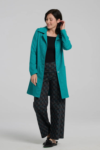 A woman wearing the Emmanuelle Coat by Kollontai in Turquoise jacquard with the Elma Pants. She is standing in front of a white background. 