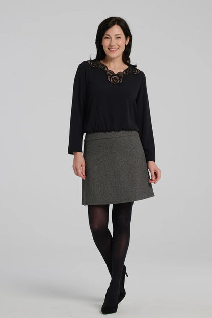 A woman wearing the Frost Skirt by Kollontai in Charcoal tweed, an above the knee length A-line skirt with an elastic waist.  She is wearing it with a black top and standing in front of a grey background. 