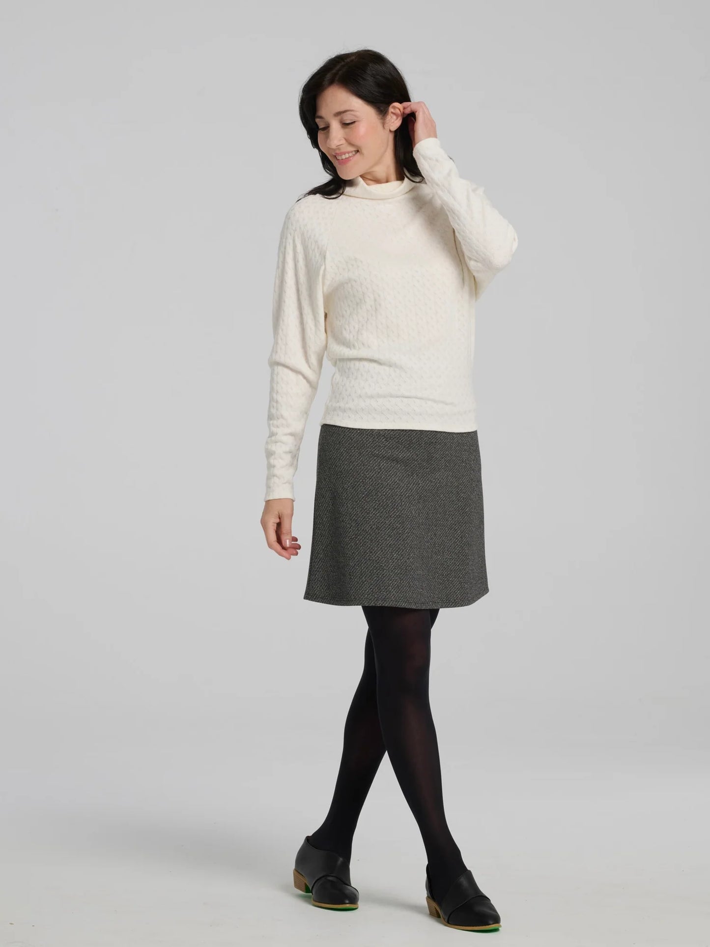 A woman wearing the Frost Skirt by Kollontai in Charcoal tweed, an above the knee length A-line skirt with an elastic waist.  She is wearing it with a black top and standing in front of a grey background. 