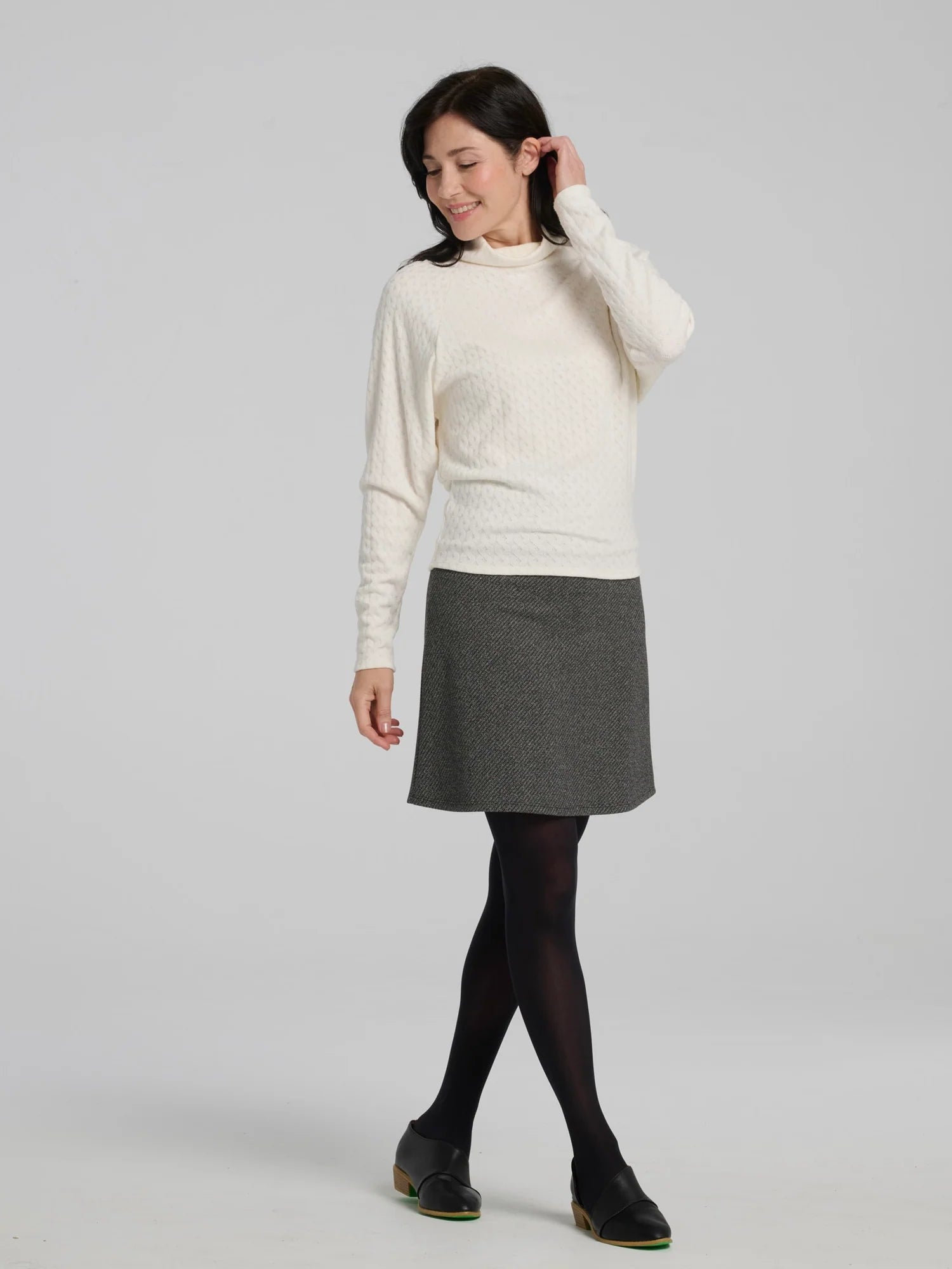 A woman wearing the Frost Skirt by Kollontai in Charcoal tweed, an above the knee length A-line skirt with an elastic waist.  She is wearing it with a black top and standing in front of a grey background. 