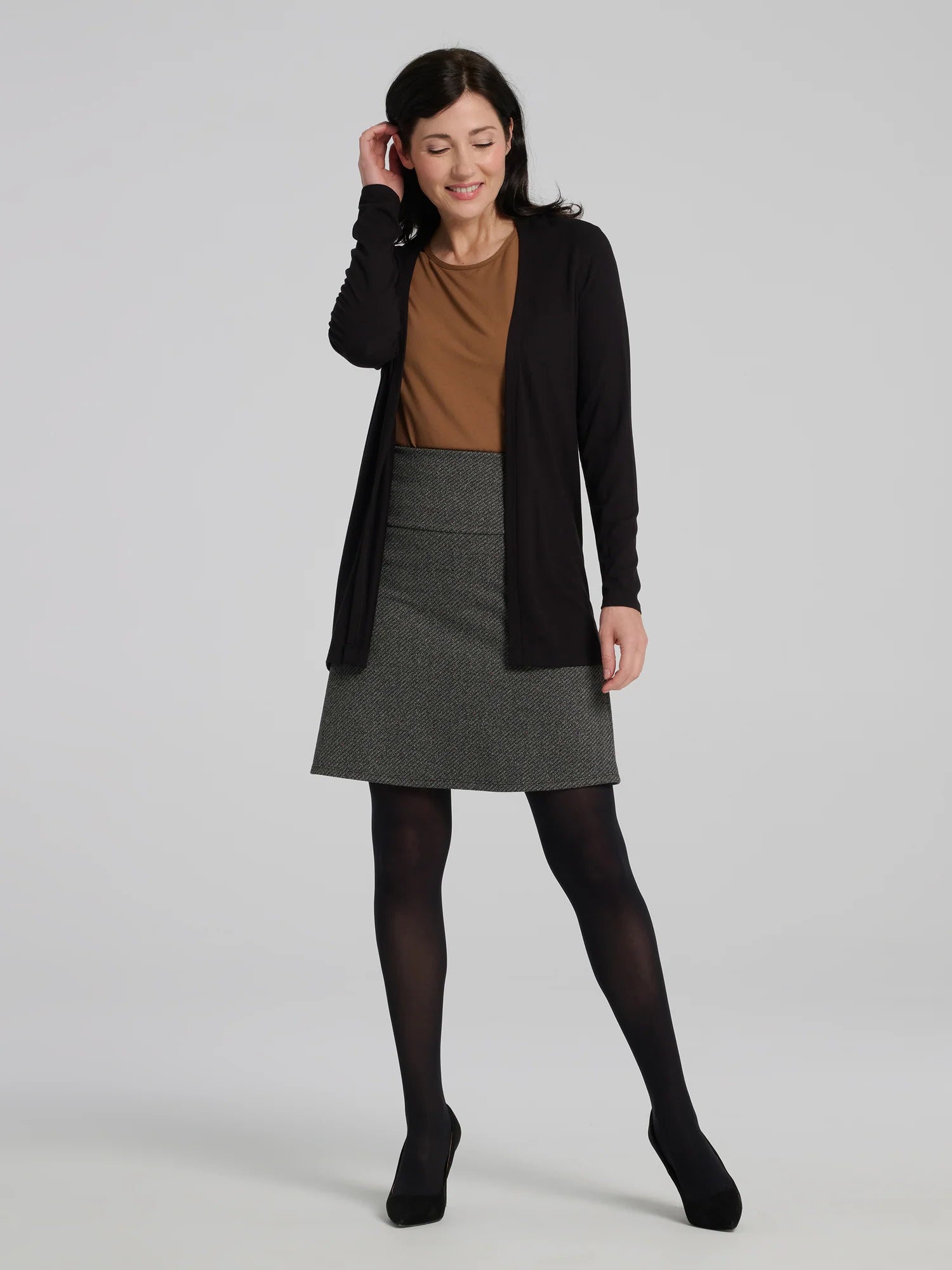 A woman wearing the Frost Skirt by Kollontai in Charcoal tweed, an above the knee length A-line skirt with an elastic waist.  She is wearing it with a brown top and black cardigan top and standing in front of a grey background. 