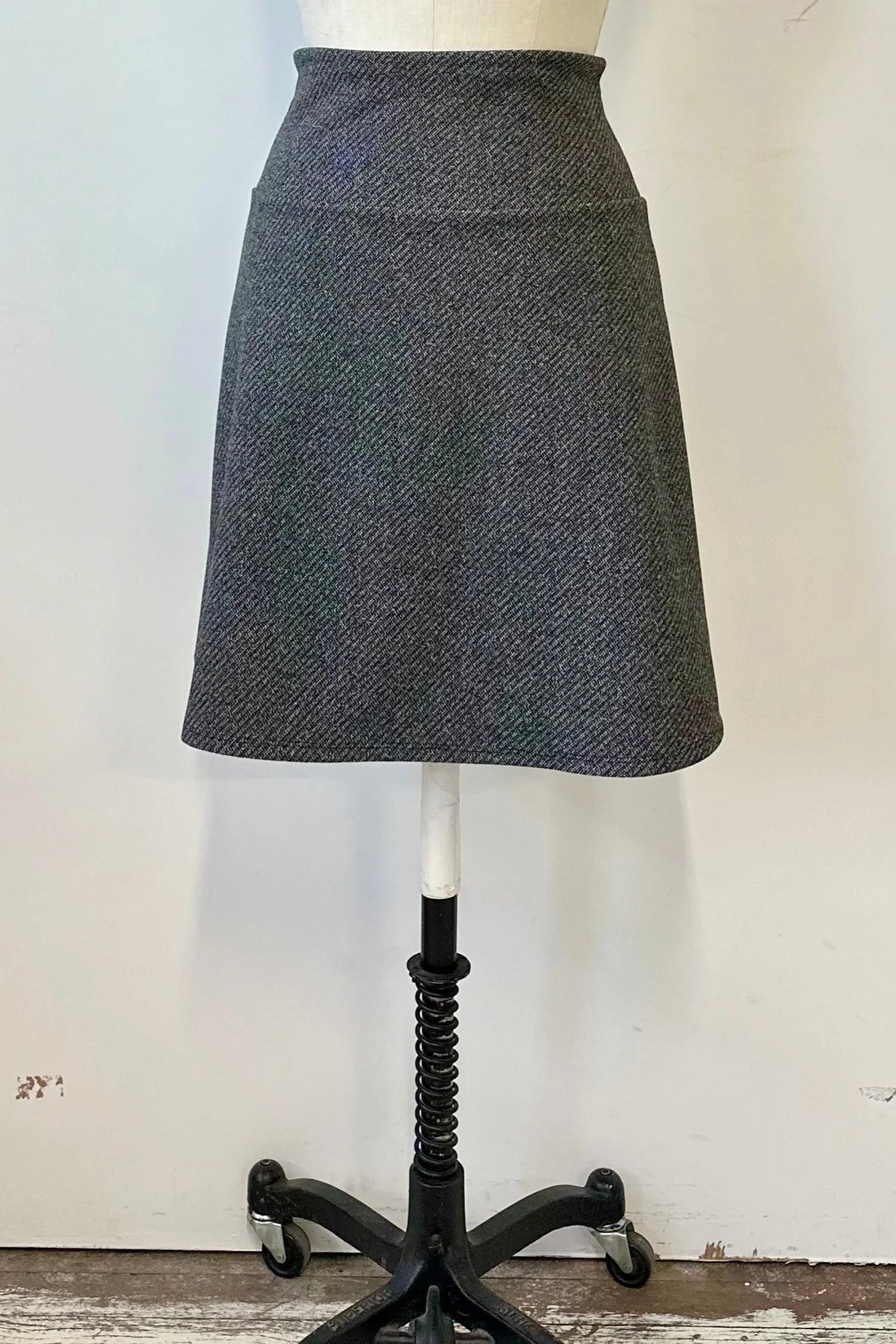 A back view of the Frost Skirt by Kollontai in Charcoal tweed, an above the knee length A-line skirt with an elastic waist, is shown on a mannequin in front of a white wall. 