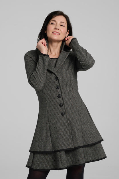 A woman wearing the Henderson Jacket by Kollontai in Charcoal tweed, with a fitted A-line shape that flares from the waist, a classic collar, and buttons down the front. She is wearing it with a matching dress and standing in front of a grey background. 