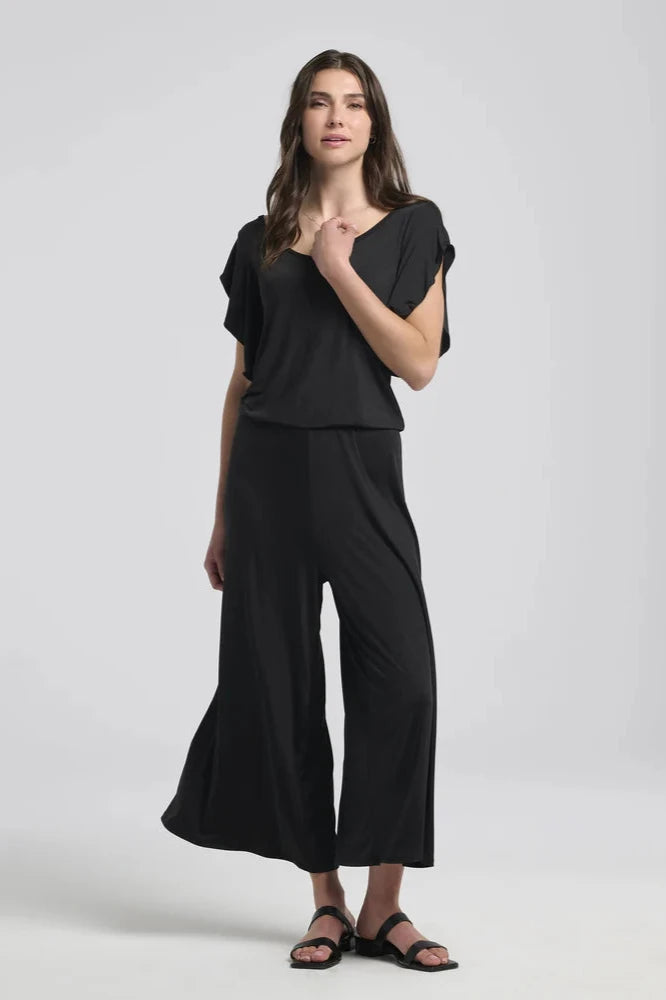 A woman wearing the Ilithyia Jumpsuit by Kollontai in Black, featuring a wide neck, short fly-away sleeves, an elastic waist, and wide 7/8 legs. She is standing in front of a white background. 