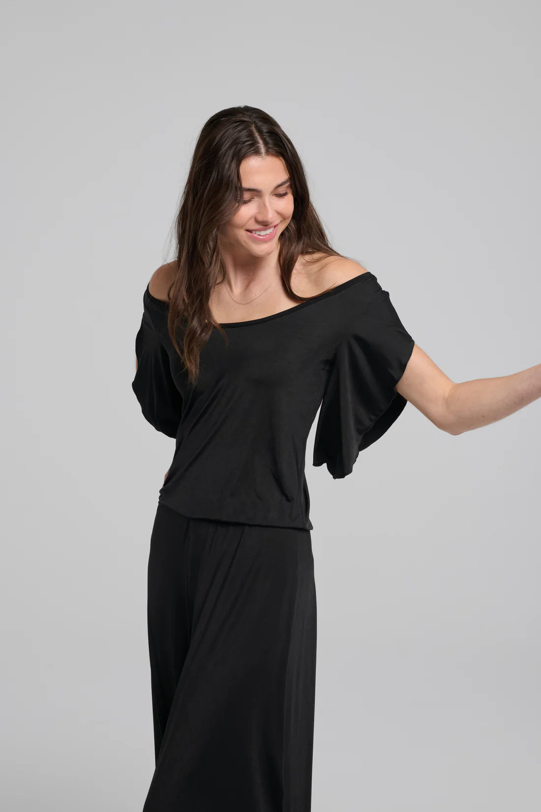 A woman wearing the Ilithyia Jumpsuit by Kollontai in Black, featuring a wide neck, short fly-away sleeves, an elastic waist, and wide 7/8 legs. She is standing in front of a white background. 