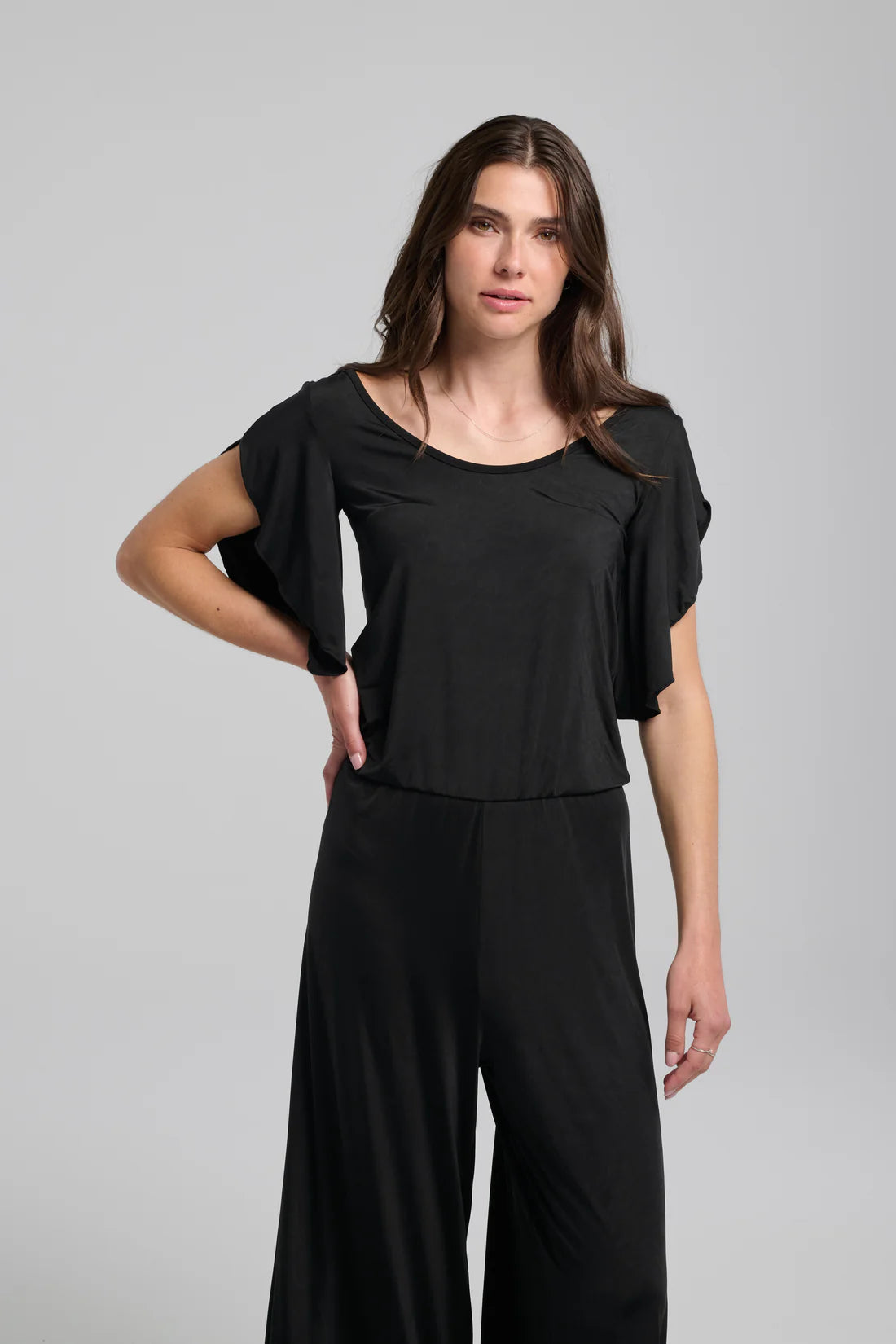 A woman wearing the Ilithyia Jumpsuit by Kollontai in Black, featuring a wide neck, short fly-away sleeves, an elastic waist, and wide 7/8 legs. She is standing in front of a white background. 