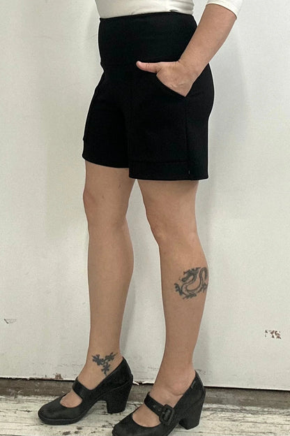 Waist down side view of a woman wearing the Milano Shorts by Kollontai, black shorts with a wide pull-one waistband and front patch pockets, standing against a white background