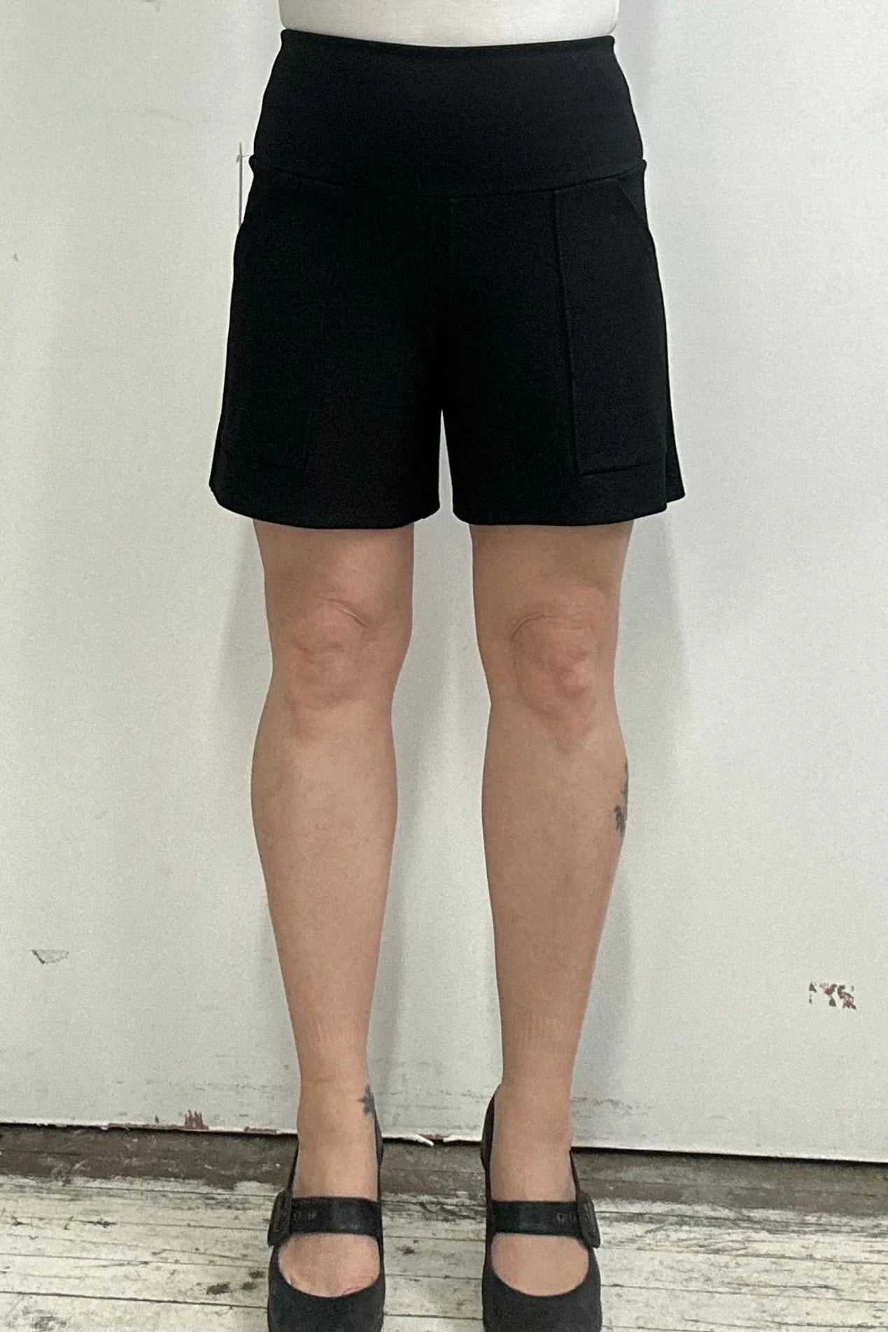 Waist down view of a woman wearing the Milano Shorts by Kollontai, black shorts with a wide pull-one waistband and front patch pockets, standing against a white background