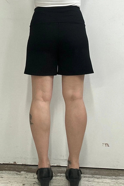 Back waist down view of a woman wearing the Milano Shorts by Kollontai, black shorts with a wide pull-one waistband and front patch pockets, standing against a white background