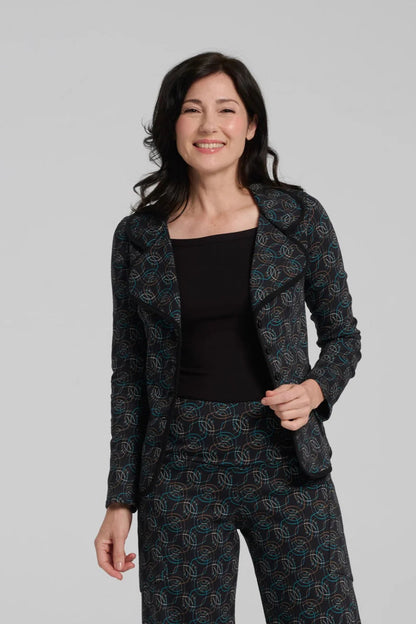 A woman wearing the Nara Jacket by Kollontai in Turquoise, a blazer with a turquoise on black geometric print, wit a rounded lapel collar, slim fit, and side pockets. She is wearing it with a black top and the matching Elma Pant and standing in front of a grey background. 