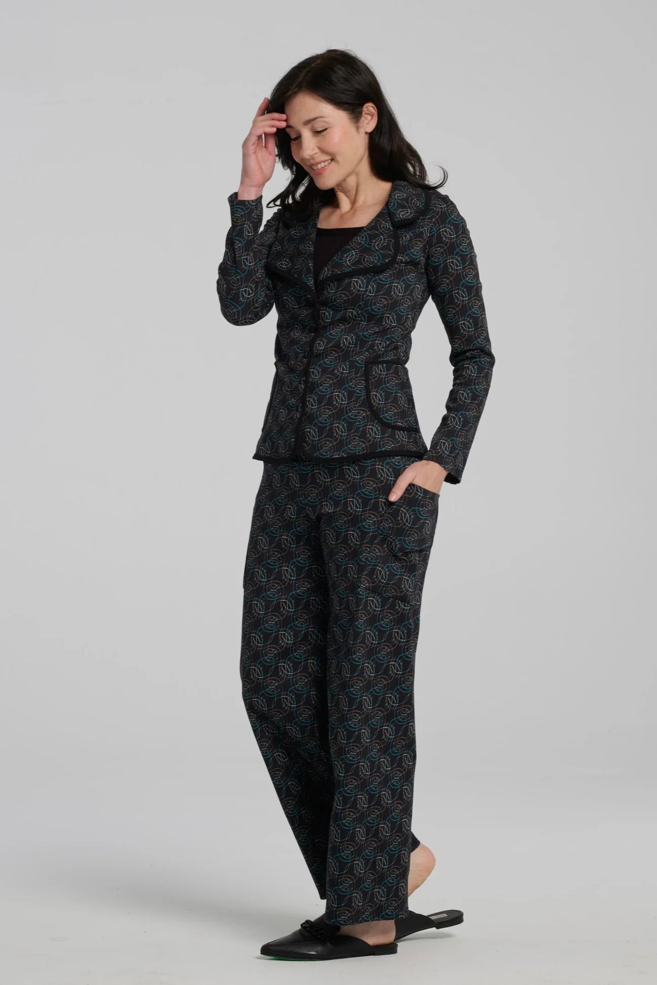 A woman wearing the Nara Jacket by Kollontai in Turquoise, a blazer with a turquoise on black geometric print, wit a rounded lapel collar, slim fit, and side pockets. She is wearing it with a black top and the matching Elma Pant and standing in front of a grey background. 