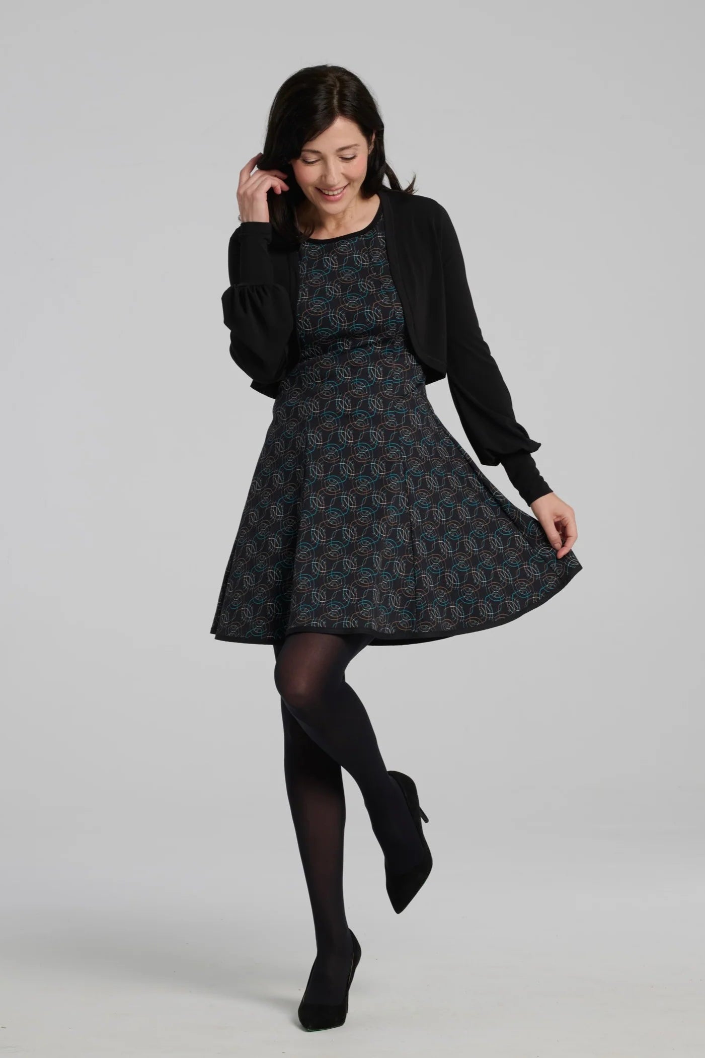 A woman wearing the Silba Dress by Kollontai in a Turquoise on black print, a sleeveless dress with a round neck, fit and flare shape, and above the knee hem. She is wearing it with a black bolero and standing against a grey background. 