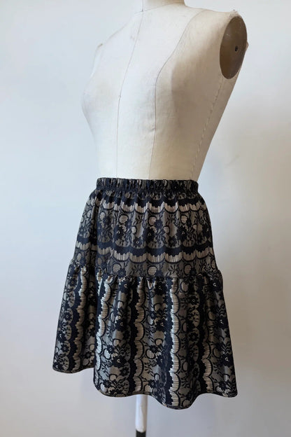 The Soleil Skirt by Kollontai, a short flounced skirt in black lace over gold satin, is shown on a mannequin  against a white background.