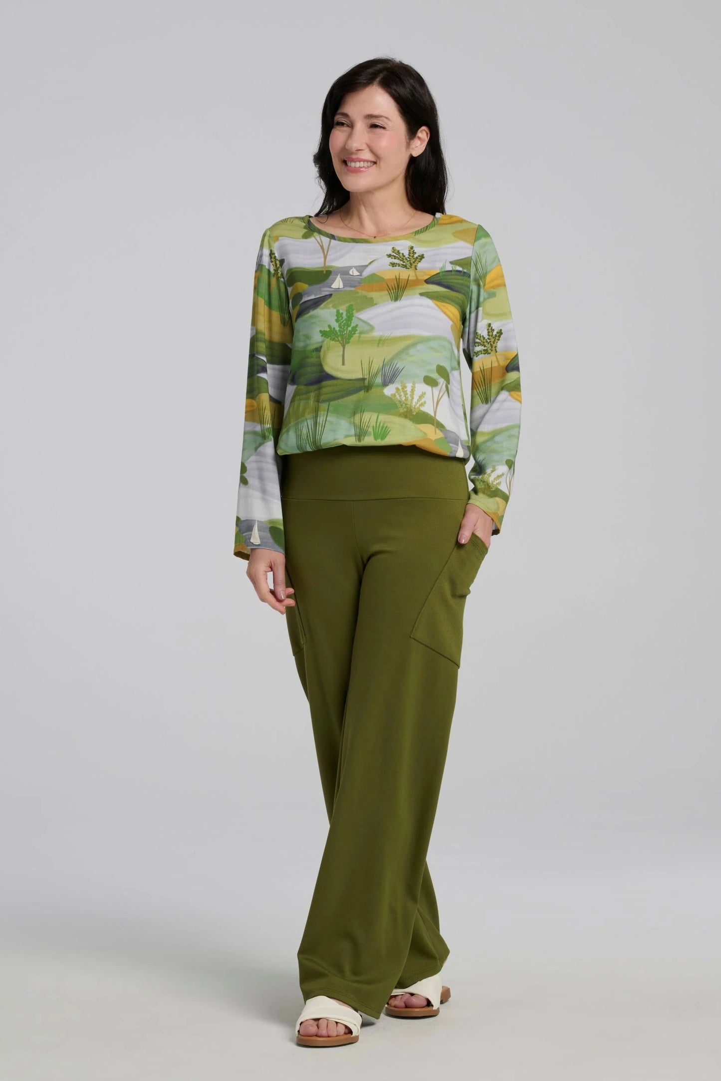 A woman wearing the spring Susan Pants by Kollontai in Granny Smith, wide leg pants with a pull-on power mesh lined waistband and angled patch pockets. She is wearing them with a patterned top and standing in front of a grey background. 
