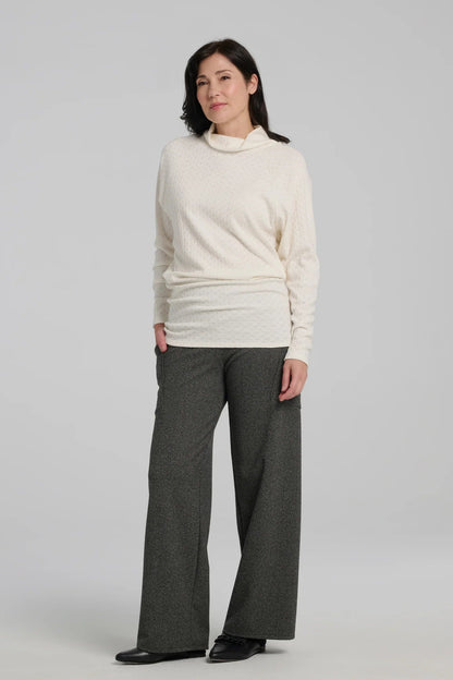 A woman wearing the Susan Pants by Kollontai in Charcoal, wide pants with a wide pull on waistband and angled patch pockets. She is wearing them with an ivory sweater and standing in front of a grey background. 