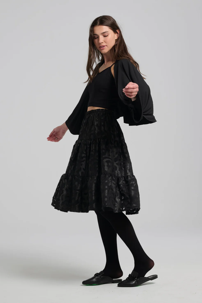 A woman wearing the Zimmer Skirt by Kollontai in Black with a black top, standing in front of a grey background