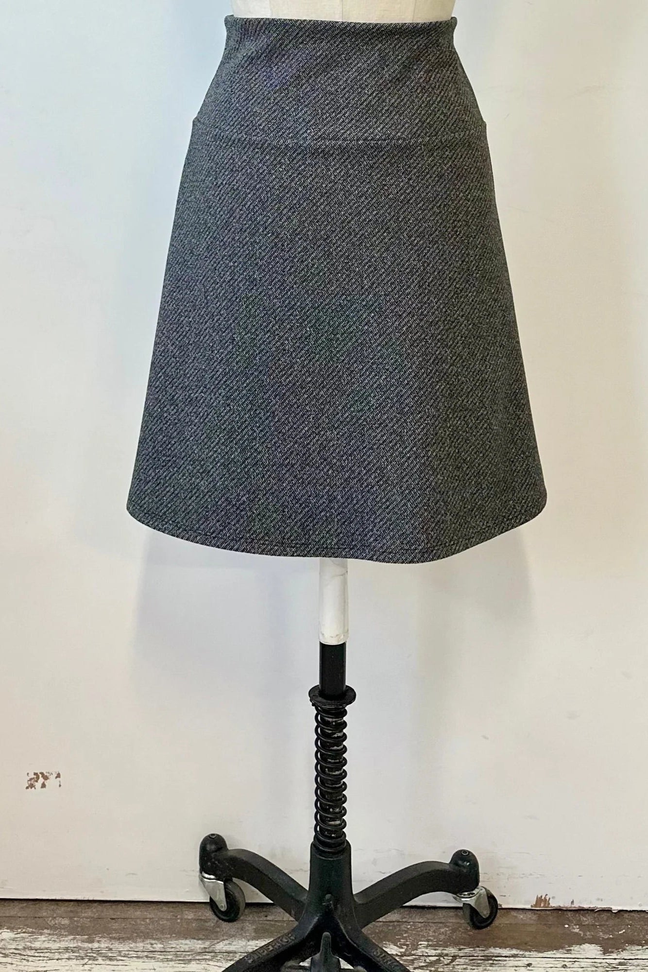 The Frost Skirt by Kollontai in Charcoal tweed, an above the knee length A-line skirt with an elastic waist, is shown on a mannequin in front of a white wall. 