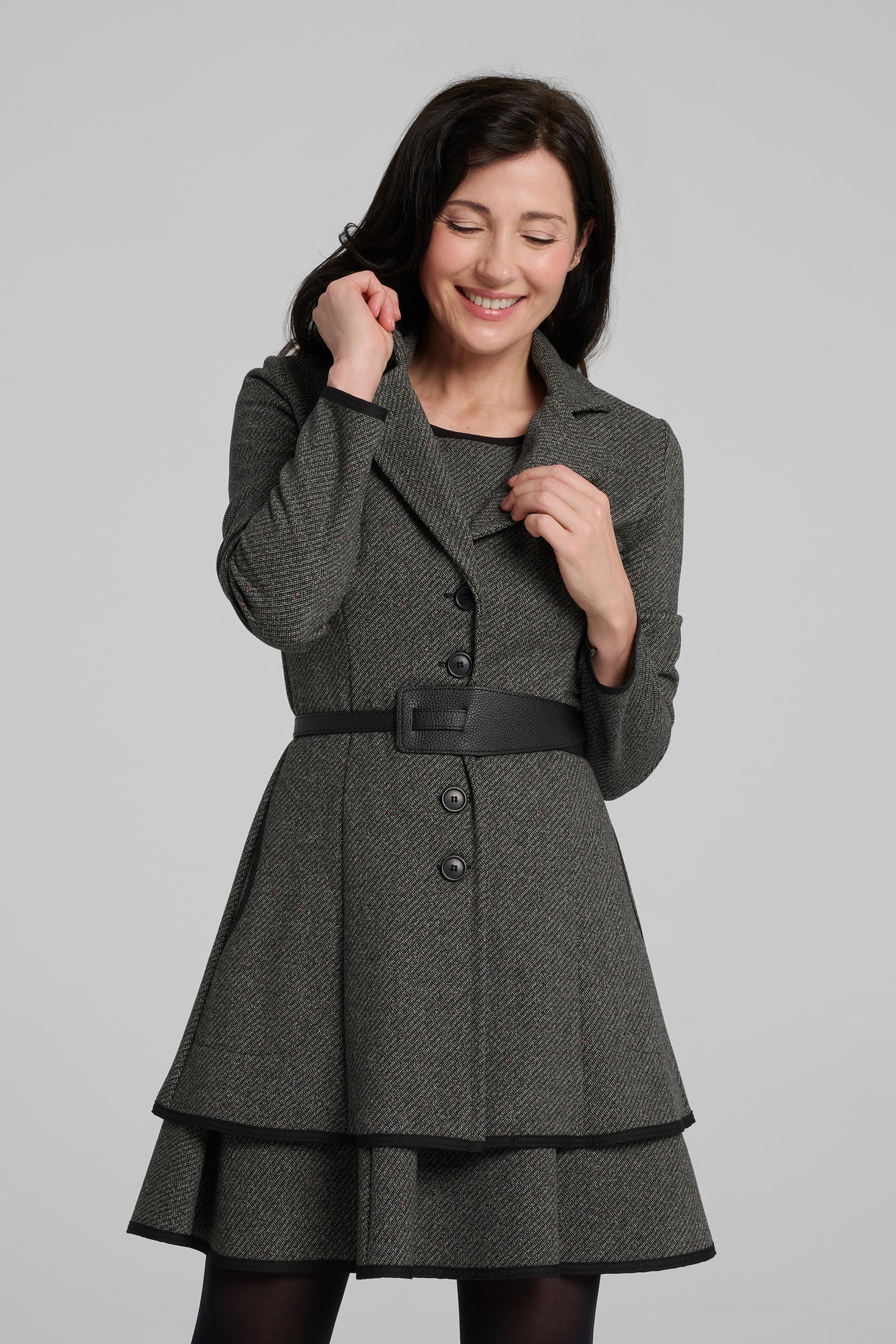 A woman wearing the Henderson Jacket by Kollontai in Charcoal tweed, with a fitted A-line shape that flares from the waist, a classic collar, and buttons down the front. She is wearing it with a matching dress and standing in front of a grey background. 