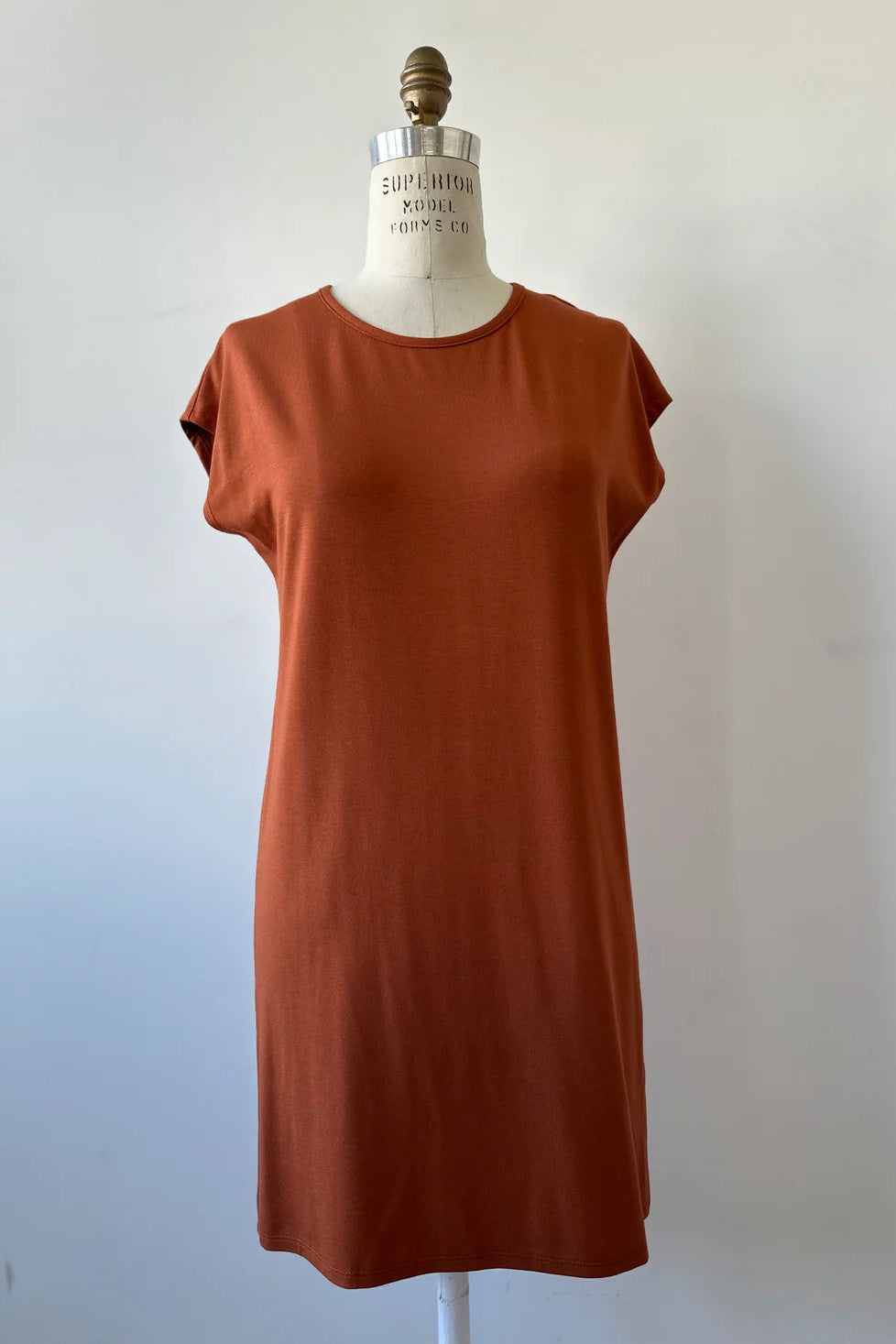 The Arwen Dress by Kollontai in Rust, a t-shirt dress with a round neck, short extended sleeves, a loose ft, and an above the knee hem, is shown on a mannequin in front of white background. 