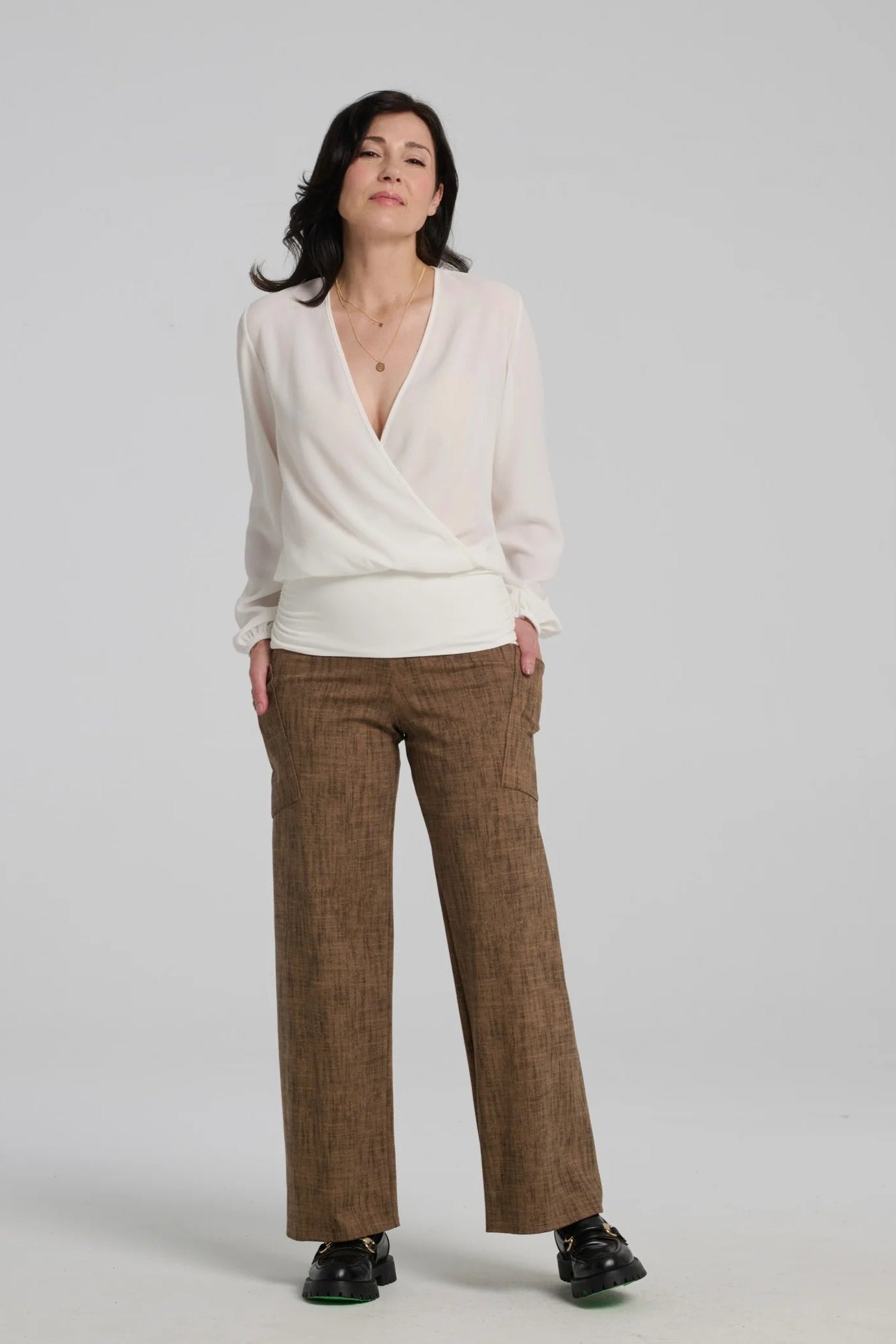A woman wearing the Susan Pants by Kollontai in Brown, wide pants with a wide pull on waistband and angled patch pockets. She is wearing them with an ivory blouse and standing in front of a grey background. 