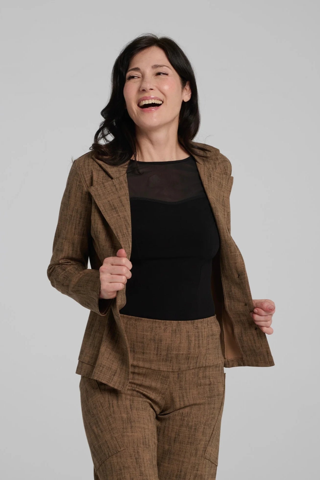 A woman wearing the Susan Pants by Kollontai in Brown, wide pants with a wide pull on waistband and angled patch pockets. She is wearing them with an black top and matching jacket and is standing in front of a grey background. 