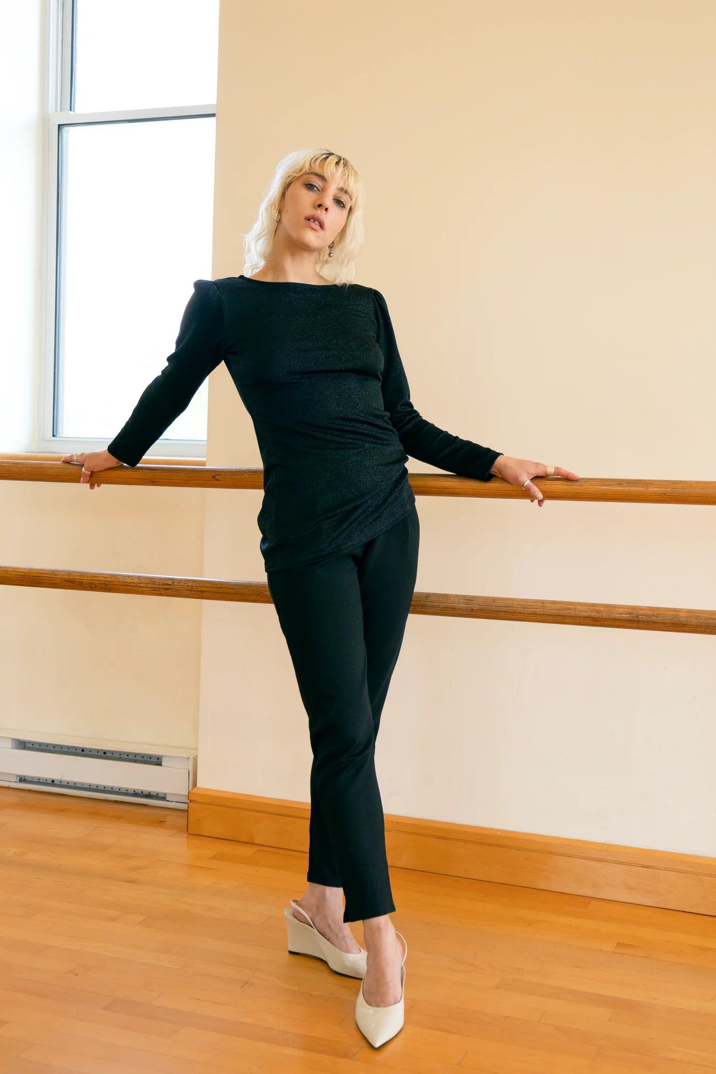 A woman wearing the Lennox Reversible Tunic by Eve Lavoie in Sparkly Black, featuring a U-neck on one side and boat neck on the other, long sleeves with puffed shoulders, and below the hip length. She is wearing it with black pants and standing in a dance studio. 