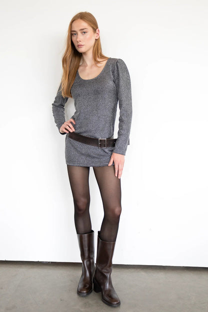 A woman wearing the Lennox Reversible Tunic by Eve Lavoie in Silver, featuring a U-neck on one side and boat neck on the other, long sleeves with puffed shoulders, and below the hip length. She is wearing it with tights and standing in front of a white wall. 