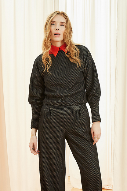 A woman wearing the Lewis Top by Melow in Black, featuring a diagonal stripe texture, boat neck, long puffed sleeves that snug at the wrists, and a cropped length. She is wearing it with matching pants and standing in front of a curtain. 