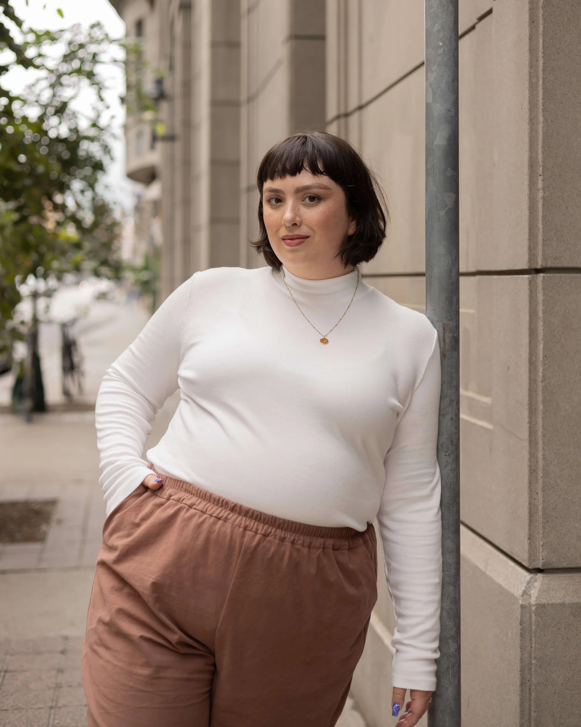A woman wearing the Essential Turtleneck by Lights of All in Cream with brown pants, standing on the street.