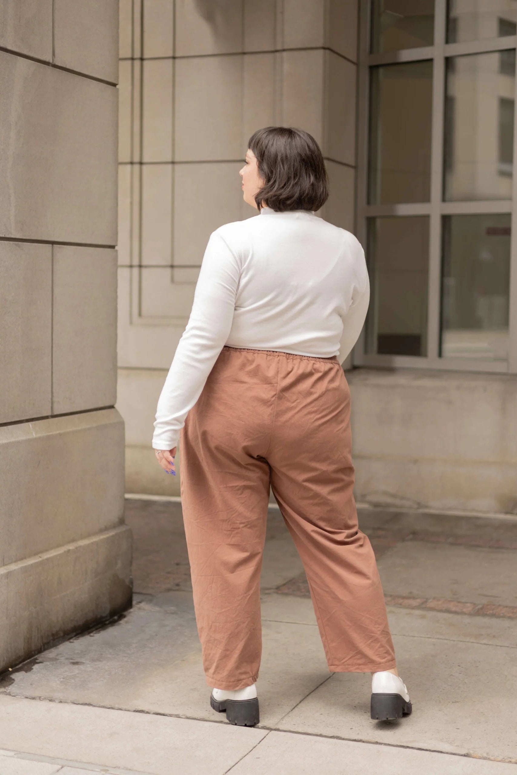 Back view of a woman wearing the Essential Turtleneck by Lights of All in Cream with brown pants, standing on the street.