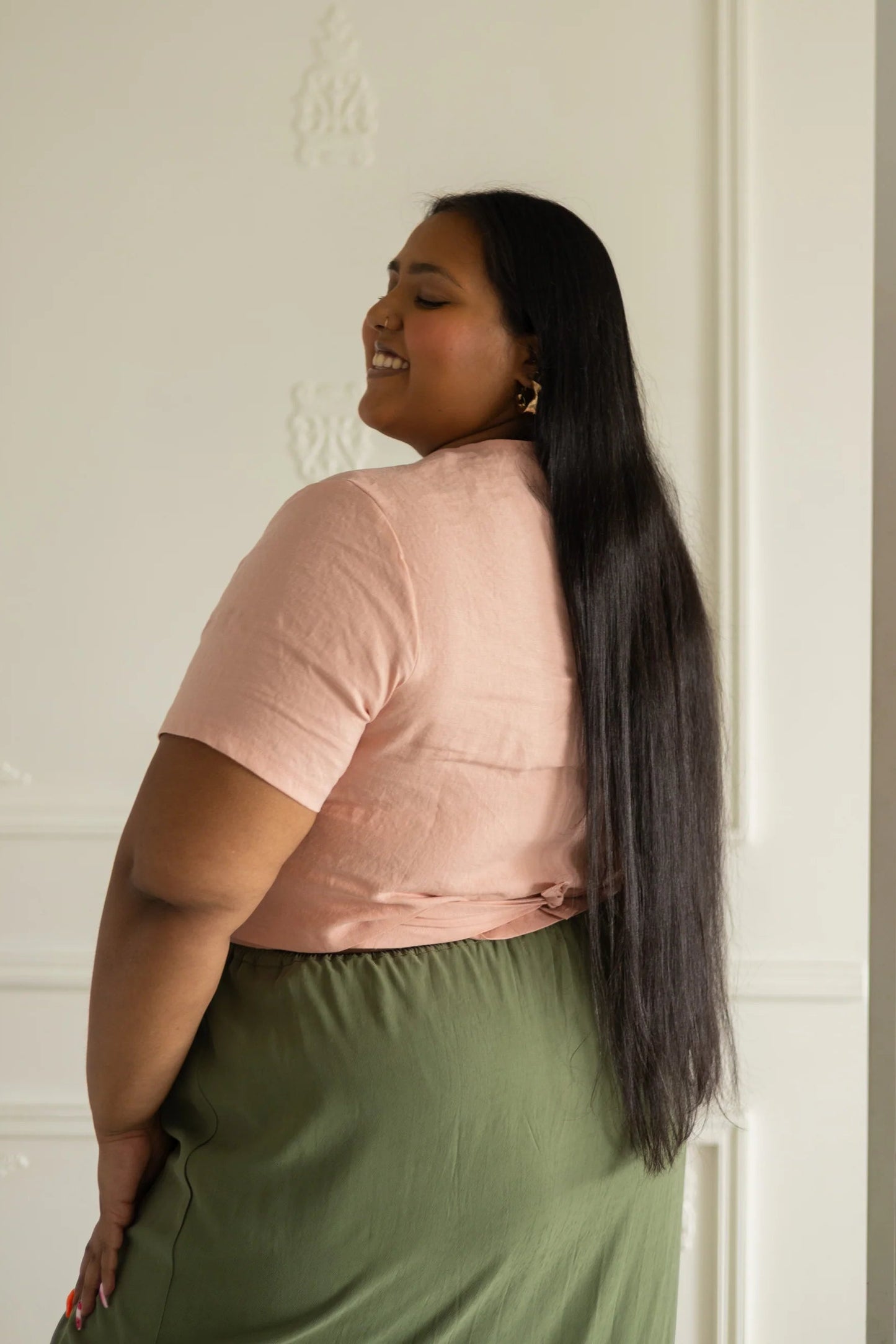 Side view of a woman wearing the Stella Top by Lights of All in Ballet Pink, a short sleeved linen wrap top that can be twisted or tied back or front. She is wearing it with the Simone Skirt in Cactus and standing in a white room. 