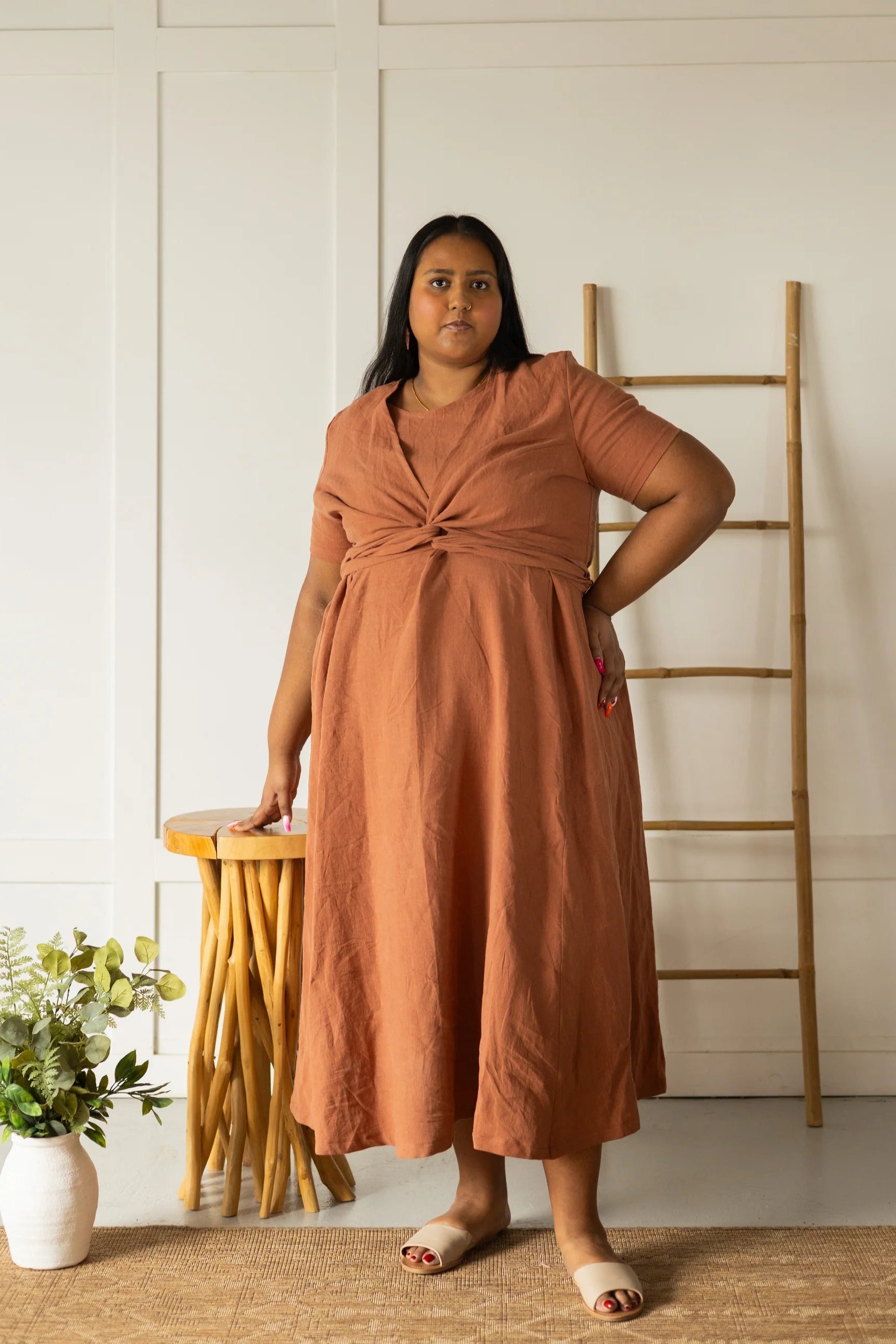 A woman wearing the Stella Top by Lights of All in Sedona, a short sleeved linen wrap top that can be twisted or tied back or front. She is wearing it over the matching Caroline dress and standing indoors by a plant stand. 
