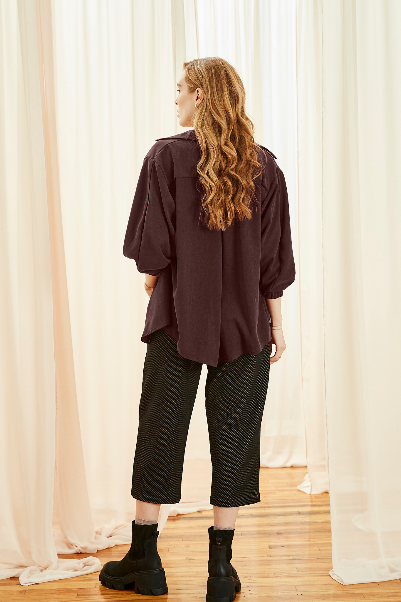 Back view of a woman wearing the Lucas Pants by Melow in Black, featuring a diagonal stripe texture, a high elastic waist, front pleats, and 3/4 length legs. She is wearing them with a burgundy shirt and standing in front of a white curtain. 