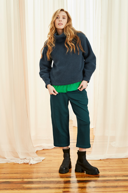 A woman wearing the Lucas Pants by Melow in Petrole (Teal/Green), featuring a diagonal stripe texture, a high elastic waist, front pleats, and 3/4 length legs. She is wearing them a green shirt and sweater and standing in front of a white curtain. 