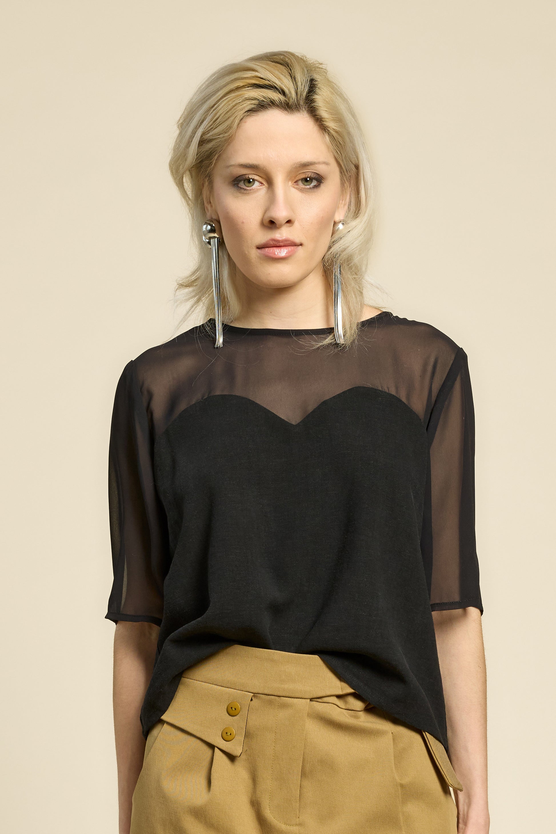 Waist up view of a woman wearing the L'Amour Top by Cokluch in Black, with a heart-shaped bodice with a sheer overlay and short sleeves. She is wearing it with beige pants against a beige background