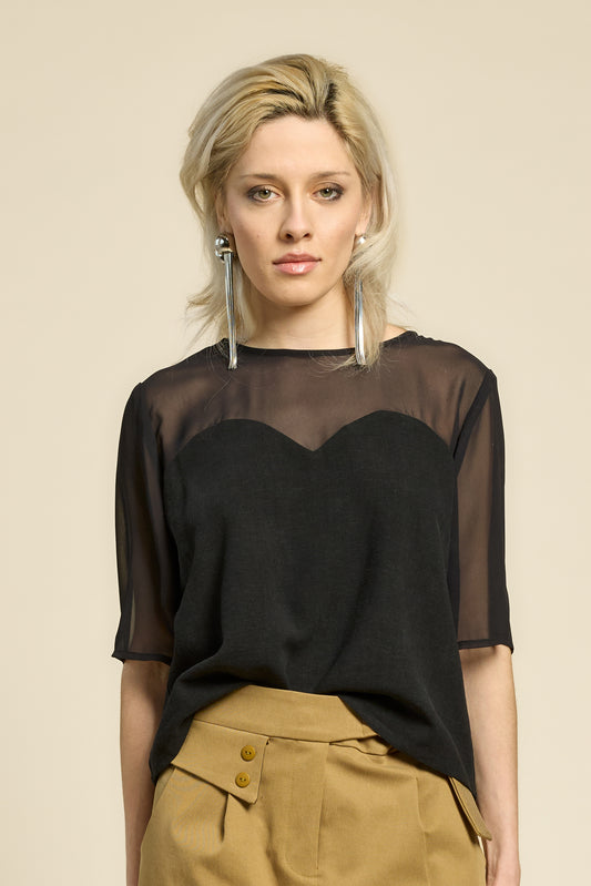 Waist up view of a woman wearing the L'Amour Top by Cokluch in Black, with a heart-shaped bodice with a sheer overlay and short sleeves. She is wearing it with beige pants against a beige background