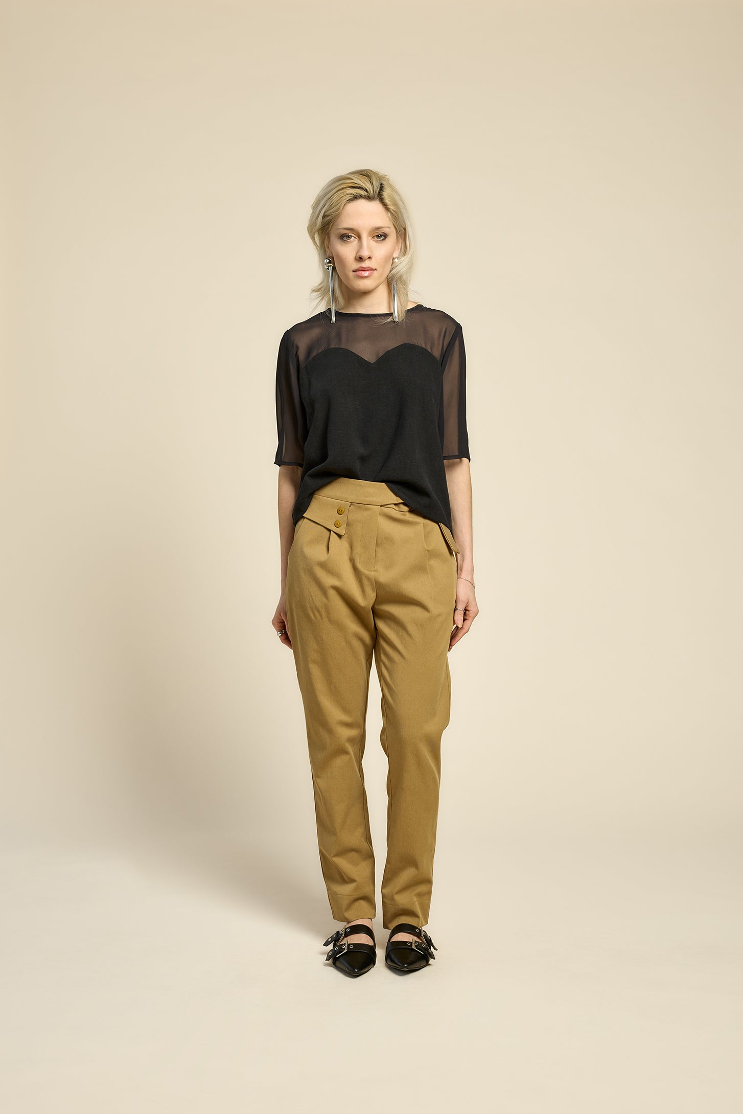A woman wearing the L'Amour Top by Cokluch in Black, with a heart-shaped bodice with a sheer overlay and short sleeves. She is wearing it with beige pants against a beige background