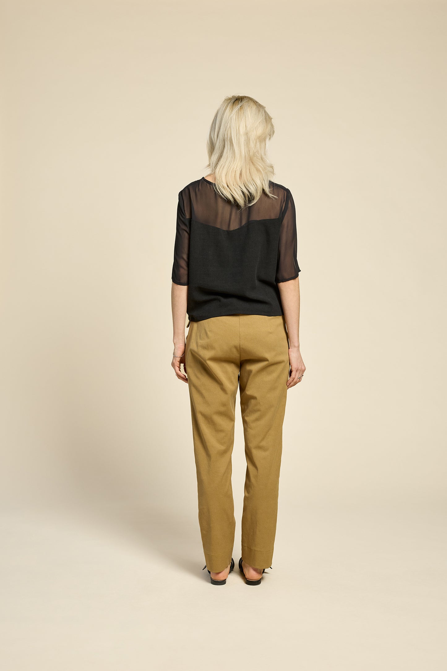 Back view of a woman wearing the L'Amour Top by Cokluch in Black, with a heart-shaped bodice with a sheer overlay and short sleeves. She is wearing it with beige pants against a beige background