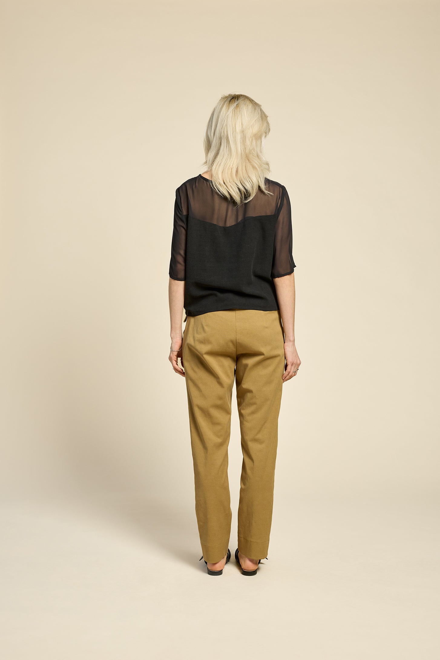 Back view of a woman wearing the L'Amour Top by Cokluch in Black, with a heart-shaped bodice with a sheer overlay and short sleeves. She is wearing it with beige pants against a beige background