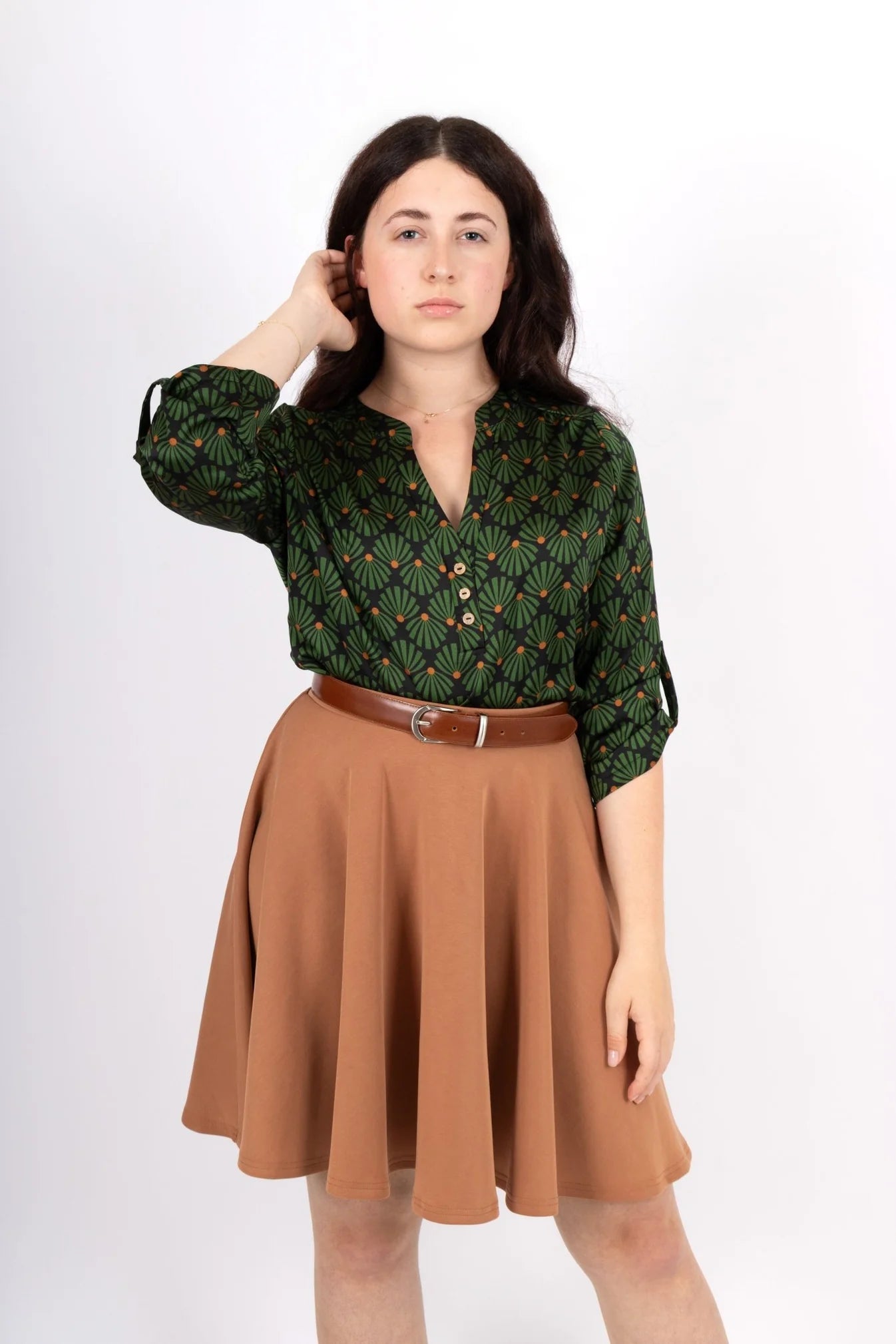 A women wearing the Leonie Blouse by Infime in Black with Green print, featuring a notched V-neckline, three button placket, and 3/4 sleeves. She is wearing it with a tan skirt and standing in front of a white background. 