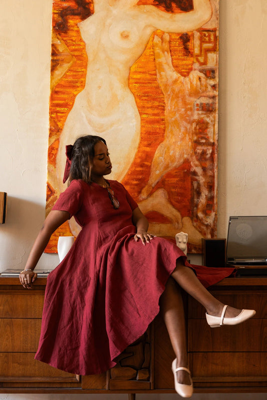 Woman wearing the Astoria Dress by Lights of All in Garnet, sitting on a wooden desk with a large painting in the background